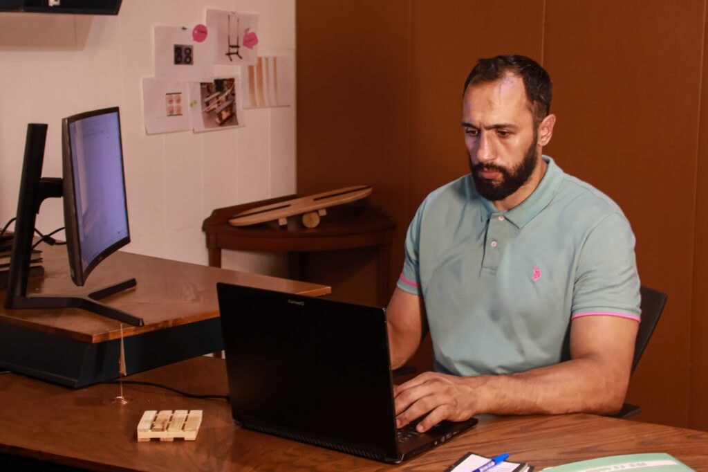 Ali Haidar working on a laptop in the HAI studio, with a balance board prototype and design screen beside him